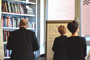 Zwei Frauen und ein Mann stehen vor einem Flipchart-Papier, das am Fenster klebt