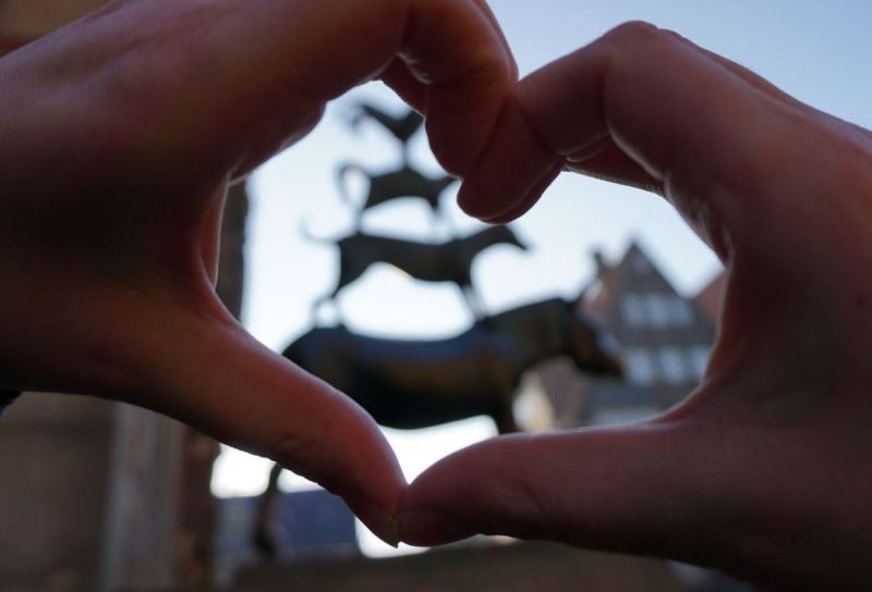 Eine hand zeigt die Form eines Herzens. Im Hintergund sind ie Bremer Statdmusiknaten als Statue zu sehen.