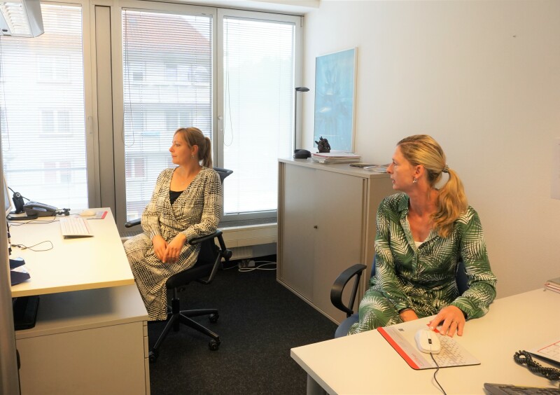 Zwei Frauen sitzen in einem Büro an zwei Tischen und schauen nach links