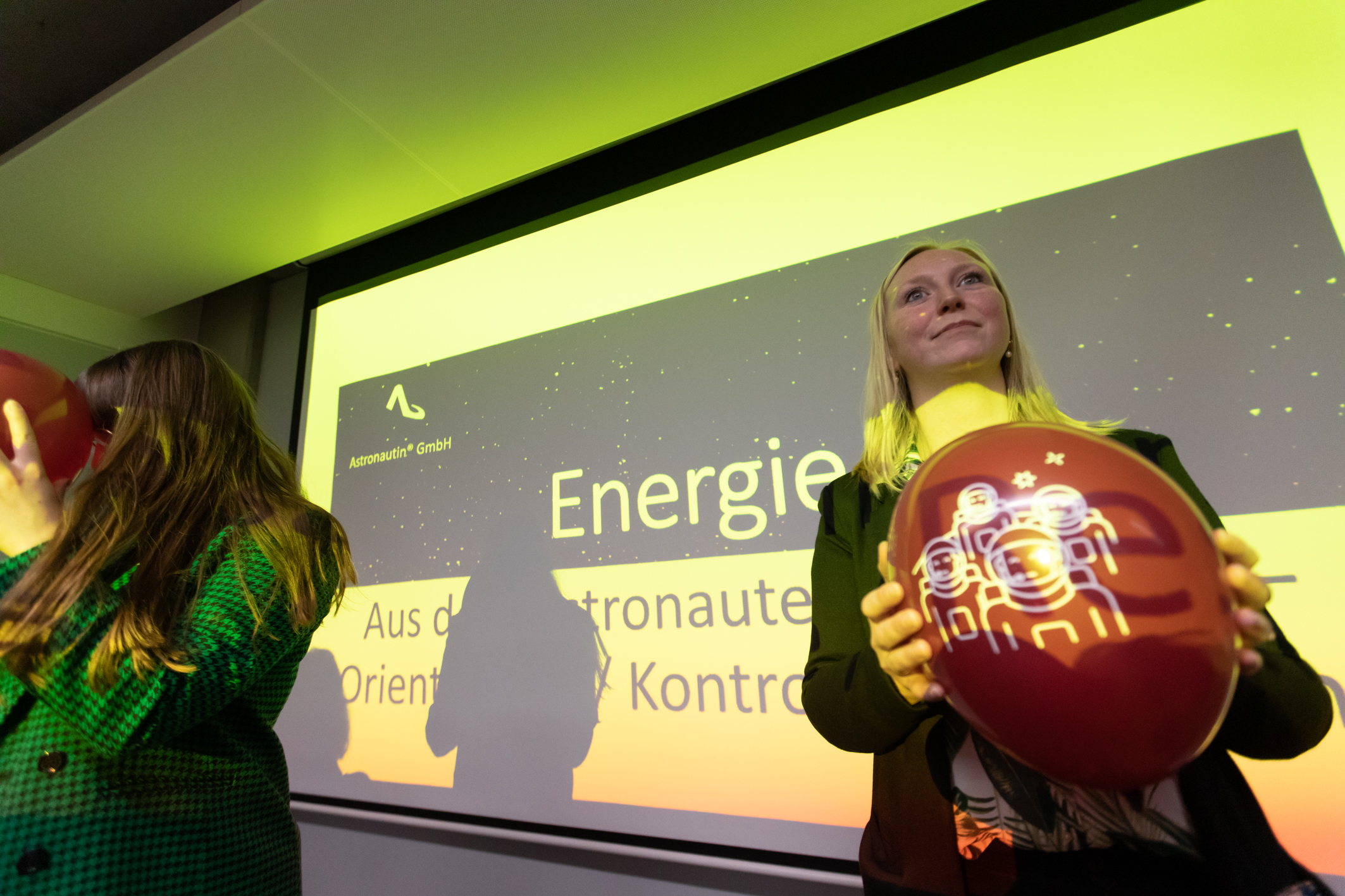 Zwei Frauen stehen vor einer Wand auf der eine Präsentation läuft, eine von Ihnen hält einen Ballon