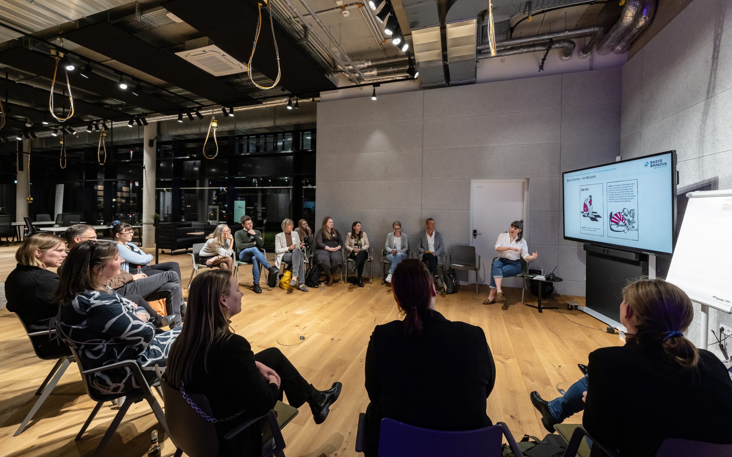 Teilnehmende eines Workshops sitzen in einem Stuhlkreis und schauen eine Präsentation auf einem Monitor an